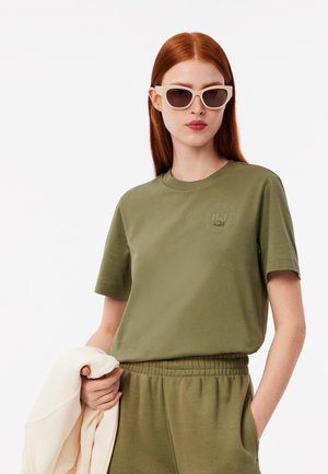 Olive green cotton t-shirt with short sleeves and a round neck, featuring a subtle logo printed in the center. Pair of white sunglasses worn.