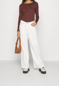 Brun langærmet top, hvide vide jeans, beige platform sko med sorte såler og en brun håndtaske med et buet håndtag.