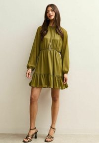 Vestido verde oliva de manga larga, cintura ceñida y bajo con volantes. Tela ligera con una textura suave. Combinado con tacones negros de tiras.