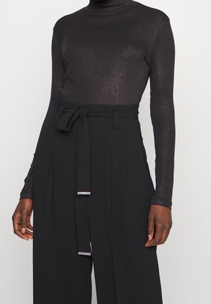 Personne portant un col roulé noir scintillant à manches longues, rentré dans un pantalon noir taille haute à jambes larges avec une ceinture nouée.