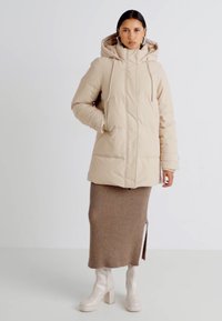 Parka matelassée beige avec capuche, dotée de poches frontales et d'une coupe droite. Associée à une jupe marron côtelée et des bottes beiges jusqu'aux genoux.