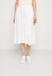 Falda midi de algodón blanco con dobladillo acampanado, cierre frontal de botones y sutil textura vertical. Combinada con zapatos blancos sin cordones.