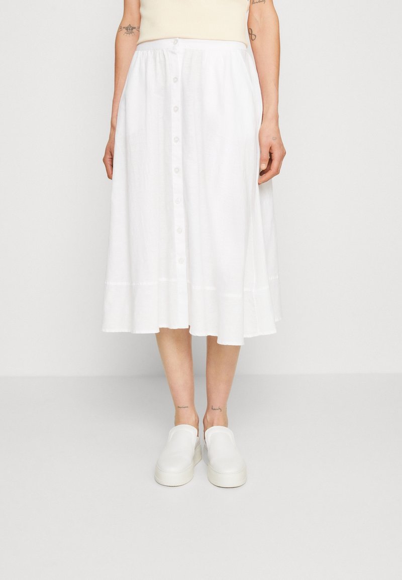 Falda midi de algodón blanco con dobladillo acampanado, cierre frontal de botones y sutil textura vertical. Combinada con zapatos blancos sin cordones.