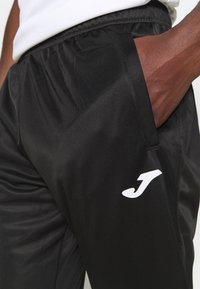 Zwarte sportbroek met een gladde textuur, elastische tailleband en wit logo aan de zijkant. Voorzien van zijzakken voor praktisch gebruik.