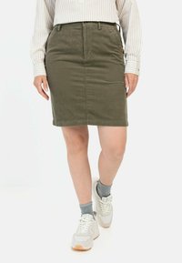 Olijfgroene corduroy rok met een aansluitend ontwerp, voorzakken en een lengte tot halverwege de bovenbenen, gecombineerd met lichtgekleurde sneakers en enkelsokken.
