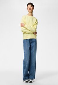 Persoon met kort haar die een lichtgele trui, wijde blauwe spijkerbroek en witte sneakers draagt, staand met gekruiste armen.