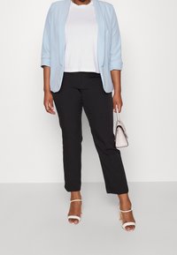 Woman wearing light blue blazer, white top, black pants, white heels, holding small pale pink handbag against plain background.