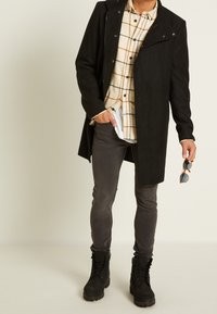 Musta mokka takki korkealla kauluksella, beige ruudullinen paita, harmaat farkut ja mustat maiharikengät. Pitelee oransseja aurinkolaseja.