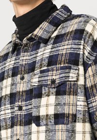 Flanellskjorta med rutmönster i marinblått, svart och krämfärgat med beige detaljer. Svarta knappar och en bröstfickdetalj är synliga.