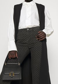 Chaleco negro sobre camisa blanca, jeans oscuros con estampado, sosteniendo un pequeño bolso negro con herrajes dorados. Tejido texturizado y diseño estructurado.