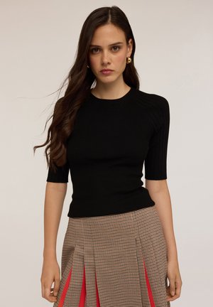 Jonge vrouw met lang bruin haar, die gouden ring oorbellen draagt, een zwart ribbelig topje met korte mouwen en een geruite rok met rode plooien.