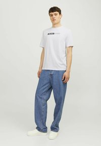 Witte katoenen t-shirt met een zwart grafisch logo. Gecombineerd met blauwe losse denim jeans en witte sneakers. Eenvoudige achtergrond.
