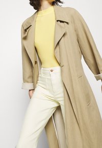 Beige linned trenchcoat med store revers, knaplukning og sidelommer, lagd over en gul tætsiddende top og lysegule højtaljede bukser.