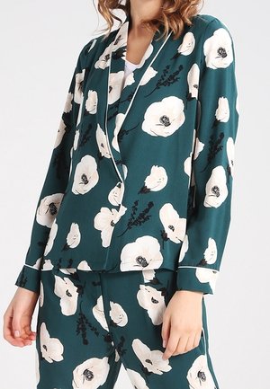 Vrouw die een donker groen bijpassend pak draagt met een grote witte bloemenprint en witte biezen langs kraag en manchetten.