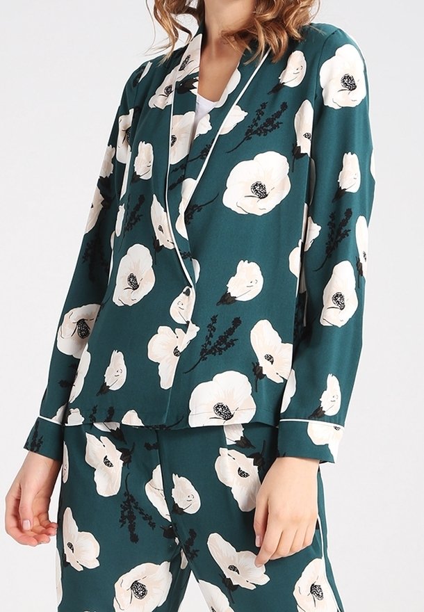 Femme portant un costume assorti vert foncé avec un grand imprimé de fleurs blanches et un liseré blanc le long du col et des poignets.