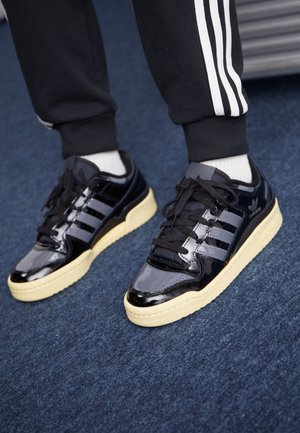 Personne portant des baskets Adidas noires et grises avec des semelles crème, des chaussettes blanches et un pantalon de survêtement noir avec des bandes blanches sur un tapis bleu.