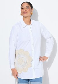 Witte blouse met lange mouwen en een knoopsluiting, met een groot beige haakpatroon aan de linkerkant. Gladde stof, met kraagontwerp.