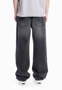 Oseba, oblečena v široke črne kavbojke in svetlo sivo majico, stoji z obrazom obrnjenim proč od kamere na belem ozadju.