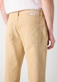 Pantaloni din bumbac bej cu croială dreaptă, având două buzunare la spate și un etichetă de marcă vizibilă pe talie. Detalii de țesătură texturată.