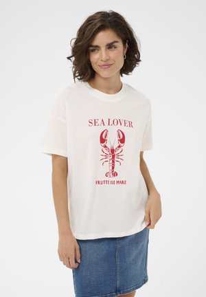 Femme aux cheveux bruns bouclés courts portant un t-shirt blanc "SEA LOVER" avec un graphique de homard rouge et une jupe en jean, debout devant un fond blanc.