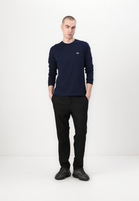 Chemise à manches longues bleu marine avec un petit logo vert, associée à un pantalon noir et des chaussures sombres, le tout sur un fond blanc uni.