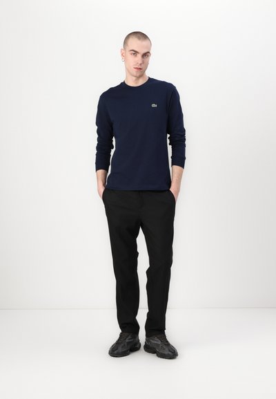 Chemise à manches longues bleu marine avec un petit logo vert, associée à un pantalon noir et des chaussures sombres, le tout sur un fond blanc uni.