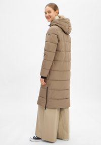 Lange beige pufferjas met een rits, verticale naden en een aangehechte capuchon. Gedragen over wijde beige broeken en zwarte sneakers.