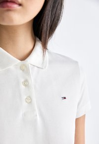 Gros plan sur une personne portant un polo blanc à trois boutons avec un petit logo rouge, blanc et bleu sur la poitrine.