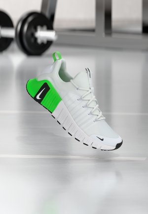 Single white and green athletic sneaker with black accents floating above a gym floor with weights blurred in background.