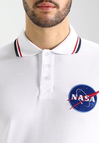 Homme portant un polo blanc avec des rayures rouges et bleu marine au col et une patch logo NASA sur la poitrine, montré de la nuque à la moitié du torse.