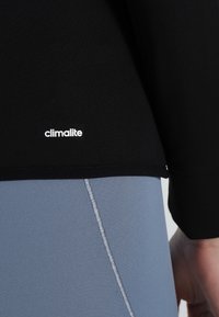 Sort træningstop med en glat overflade, med et hvidt "climalite" logo nær kanten, parret med lyseblå leggings og kontraststing.