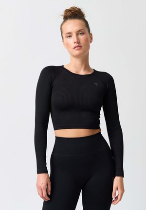 Frau mit hochgestecktem Haar, die ein schwarzes, geripptes Crop-Top mit langen Ärmeln und passende hochtaillierte Leggings vor einem einfarbigen Hintergrund trägt.
