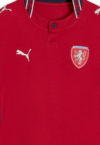 Maillot de sport rouge avec un logo Puma blanc sur la gauche et un écusson en forme de blason avec un lion argenté sur le côté droit de la poitrine.