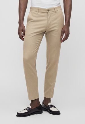 Man wearing beige slim-fit trousers and black and white loafers against a plain light background.