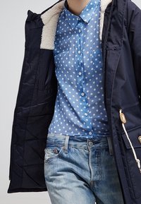 Marinblå parka med krage i pälsöverdrag över en ljusblå skjorta med prickar, kombinerat med ljusa jeans i denim. Design med knappar och synliga stygn.