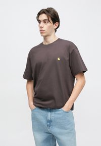 Νεαρός με σκουλαρίκι, φορώντας σκουρό καφέ μπλουζάκι Carhartt και ανοιχτούχρωμο τζιν, στέκεται με τα χέρια στις τσέπες μπροστά σε λευκό φόντο.