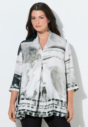 Kvinde iført løs, grå og hvid bluse med abstrakt mønster og trekvartærmer samt sorte bukser, stående mod en hvid baggrund.