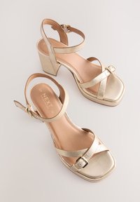 Gold metallic block-heeled sandals with ankle straps, buckle fastenings, and square open toes on a plain light background.