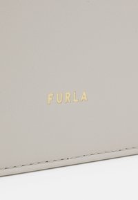 Portafoglio in pelle grigia chiara con bordo cucito e logo "FURLA" in oro a rilievo. Superficie liscia con elementi di design minimalisti.