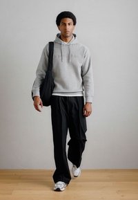 Felpa grigia con cordini, pantaloni neri morbidi e sneakers argentate. Il modello tiene in spalla una borsa nera, in piedi su un pavimento di legno.
