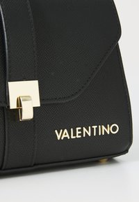 Bolso de mano de cuero negro con una superficie texturizada, que presenta un cierre cuadrado dorado y el nombre de la marca "VALENTINO" en letras doradas.