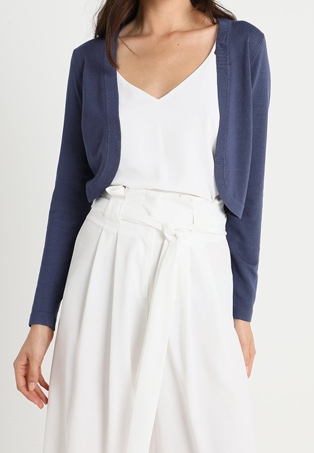Femme portant un cardigan court bleu marine sur un top blanc à col en V et un pantalon blanc taille haute à jambes larges avec une ceinture nouée.