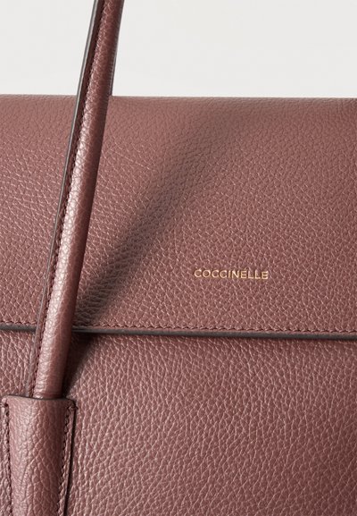Близък план на дамска чанта Coccinelle от лилаво-розова зърнеста кожа, с презрамка и златист надпис.