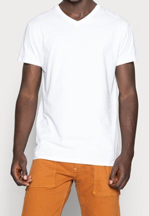 Mand iført en simpel hvid kortærmet V-hals T-shirt og orange bukser med store forlommer, stående mod en lys baggrund.