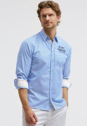 Mann trägt ein blau-weiß vertikal gestreiftes Hemd mit hochgekrempelten Ärmeln und weiße Hose vor einfachem Hintergrund.