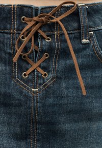 Σκούρα μπλε τζιν με μπροστινό κλείσιμο με κορδόνια από καφέ σουέντ, περασμένα από μεταλλικά κουμπώματα.