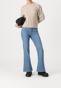 Pull beige en maille torsadée, jean flare bleu clair, et grosses bottines à plateforme noires. Pochette tissée noire. Fond minimaliste.