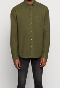 Uomo che indossa una camicia a maniche lunghe verde oliva con bottoni e tasca sul petto, abbinata a jeans neri, in piedi su uno sfondo chiaro e semplice.