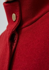 Abrigo de lana rojo con cuello alto, botón de herrajes dorados y forro interior suave. La superficie tiene un acabado texturizado y delicado.