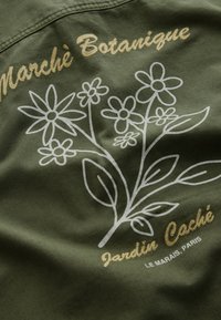 Tissu vert avec dessin floral linéaire blanc et texte cursif jaune indiquant « Marché Botanique » et « Jardin Caché Le Marais, Paris ».